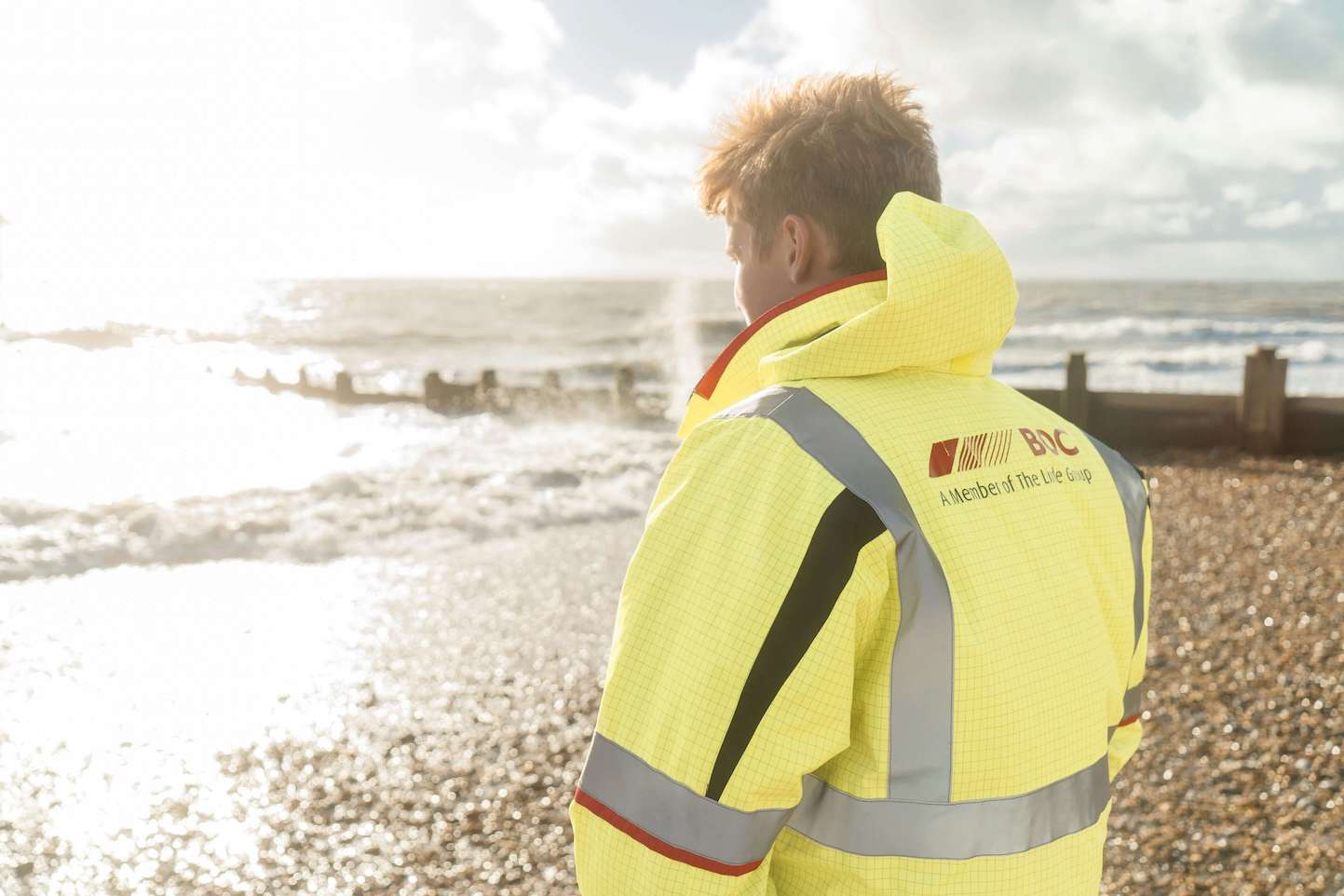 A person in a yellow high-visibility jacket stands looking at the ocean under a bright sky with sun glare, with pebbles and waves in the background.